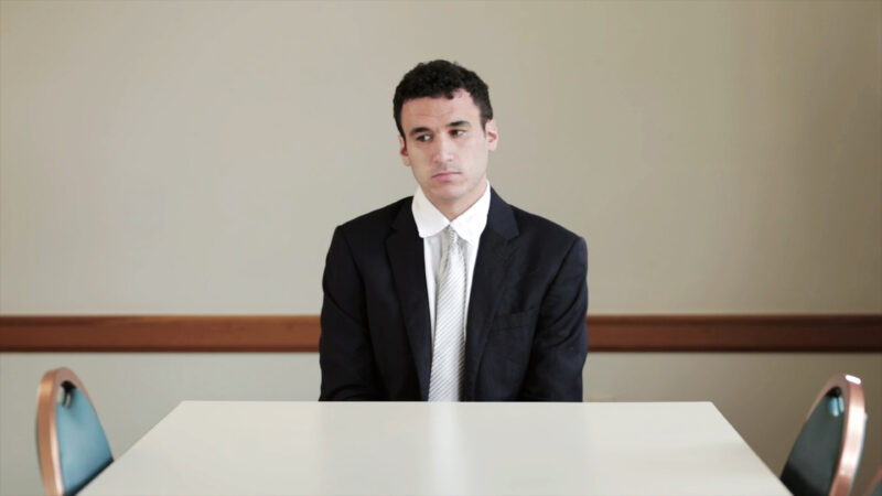 Still image from the video Mock Interview by artist Endia Beal. Image depicts a white male in suit at a table in an office setting.
